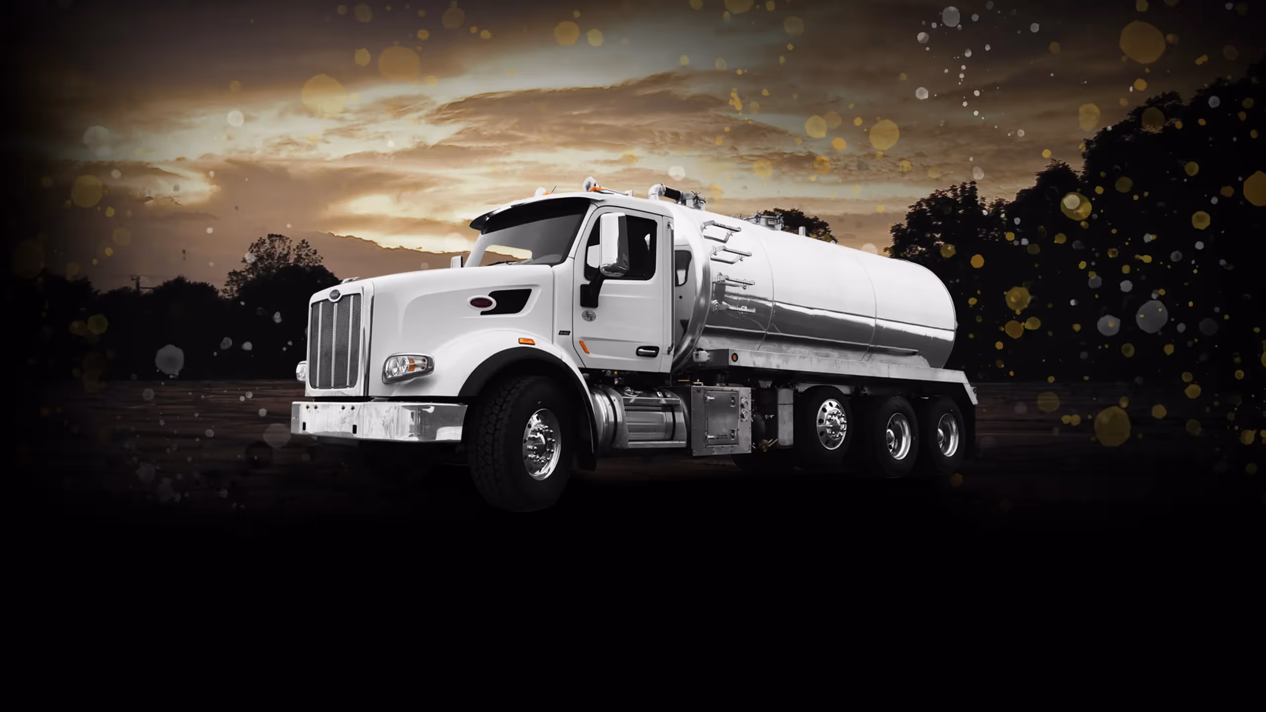White tanker truck with shiny metal tank against a dark background with a cloudy sky.