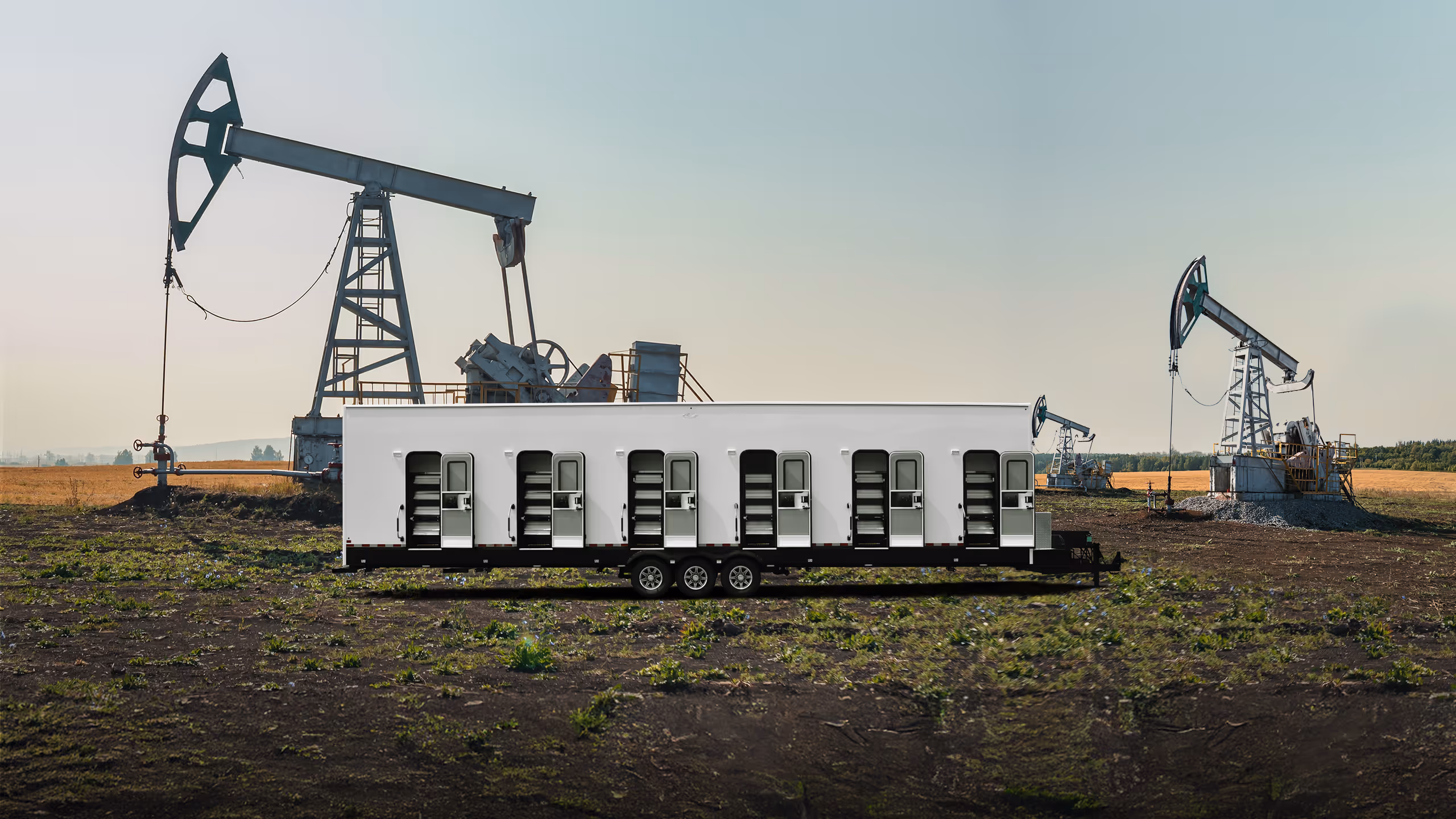 Long white mobile dormitory trailer with multiple doors set in a field with oil pump jacks.