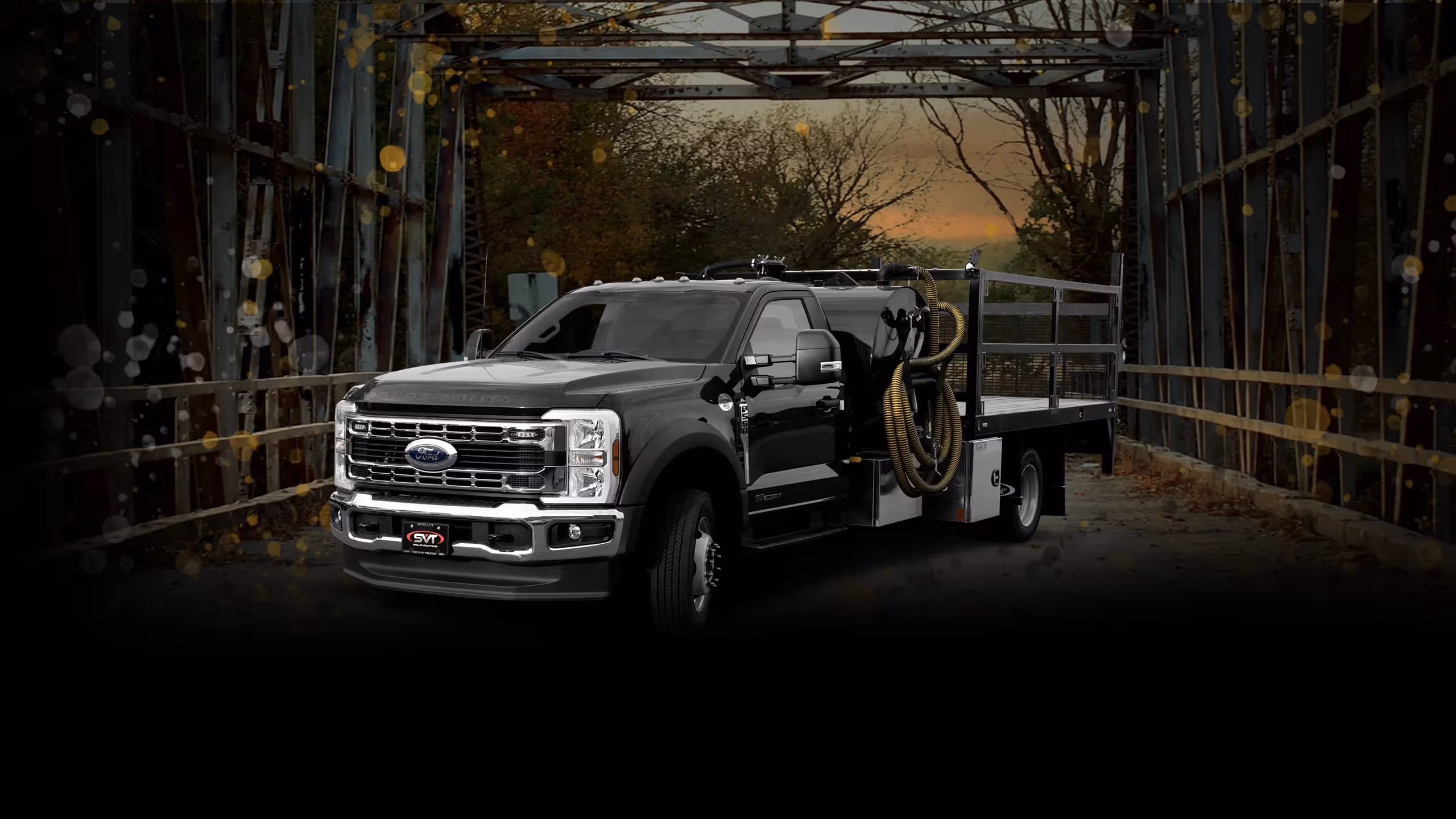Black Ford Super Duty flatbed truck with equipment parked on metal bridge at sunset.