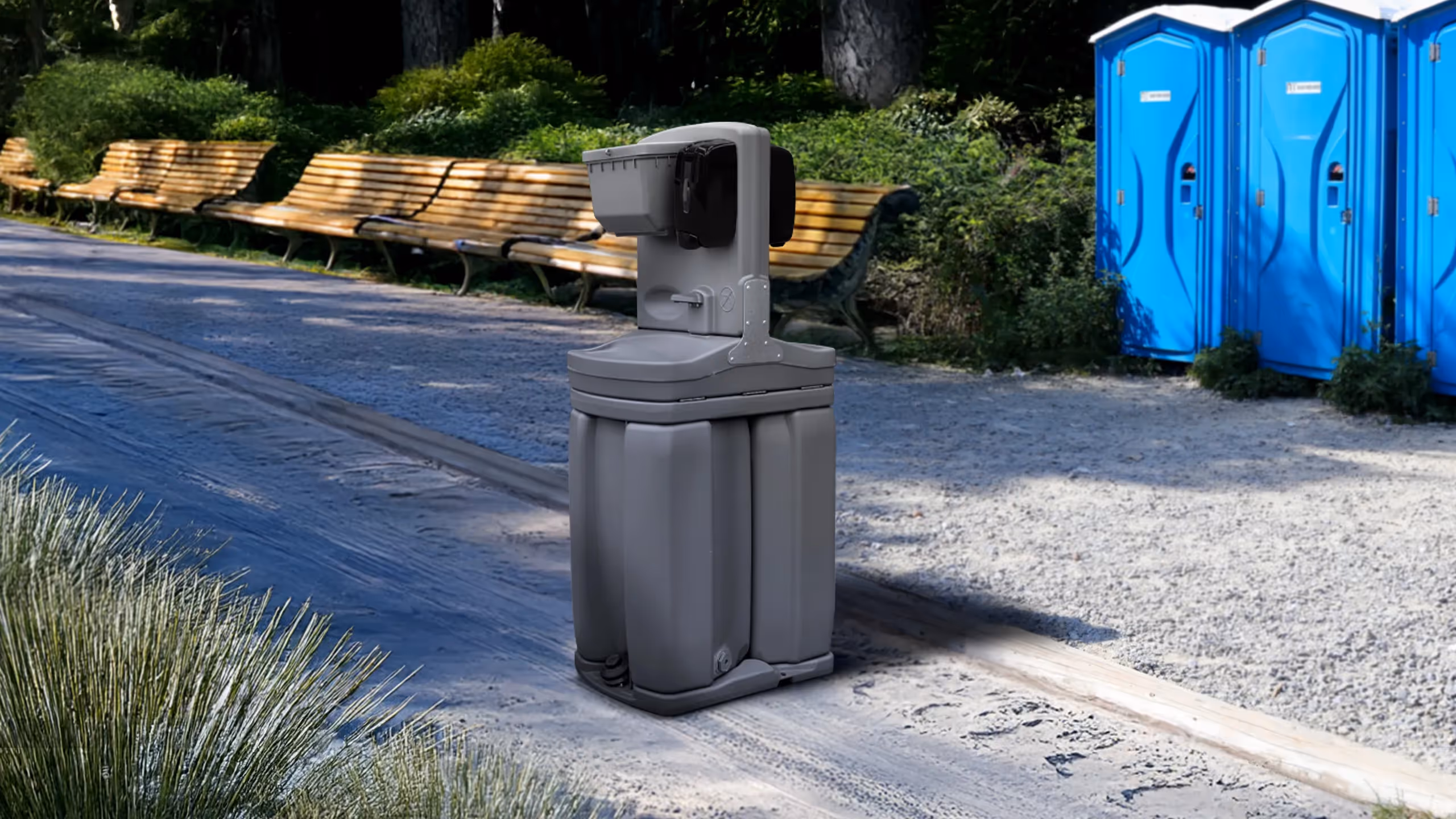 Portable handwashing station placed on a park path with benches and blue portable toilets nearby.
