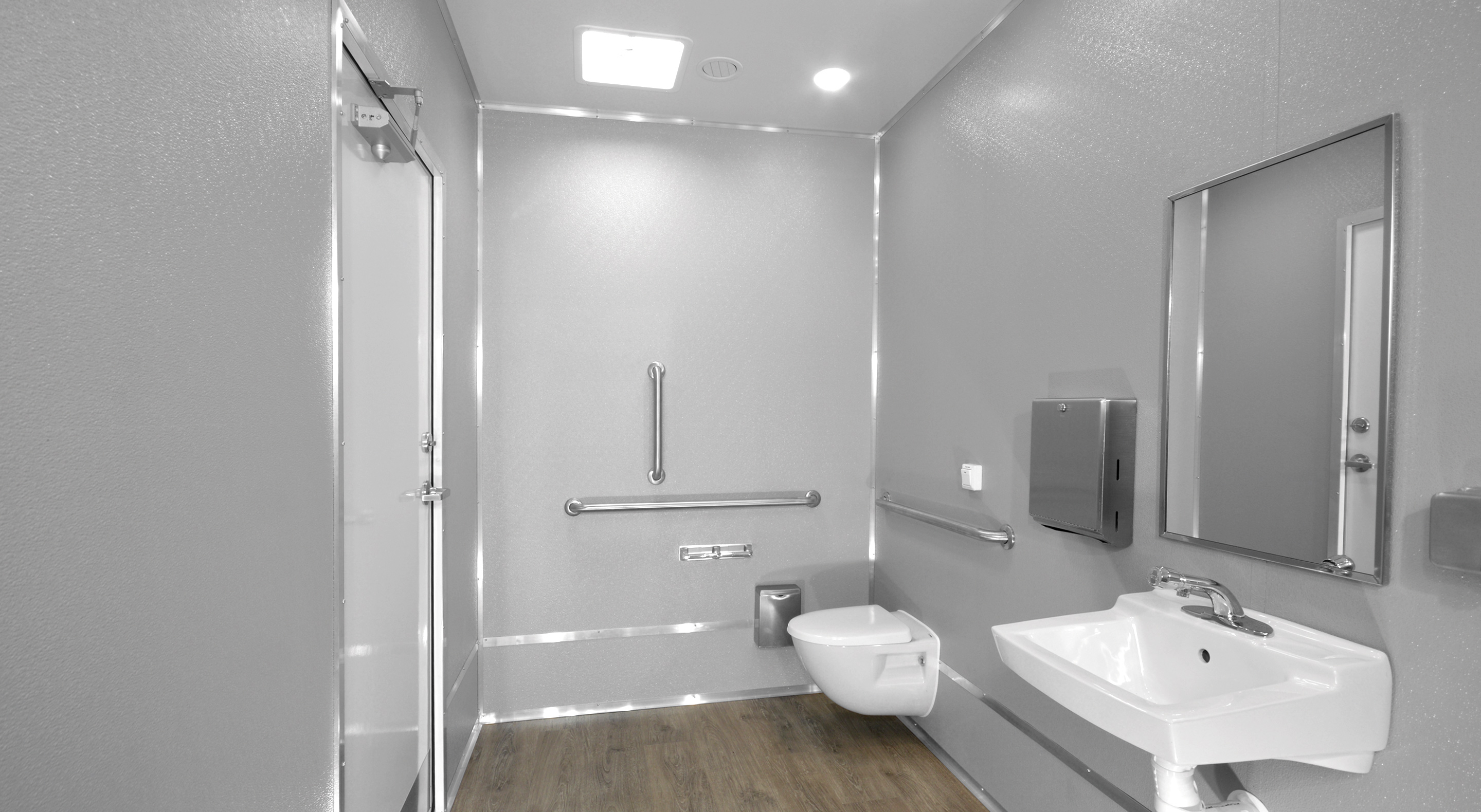 Accessible bathroom with wall-mounted toilet, grab bars, sink, mirror, hand dryer, and wood-look floor.