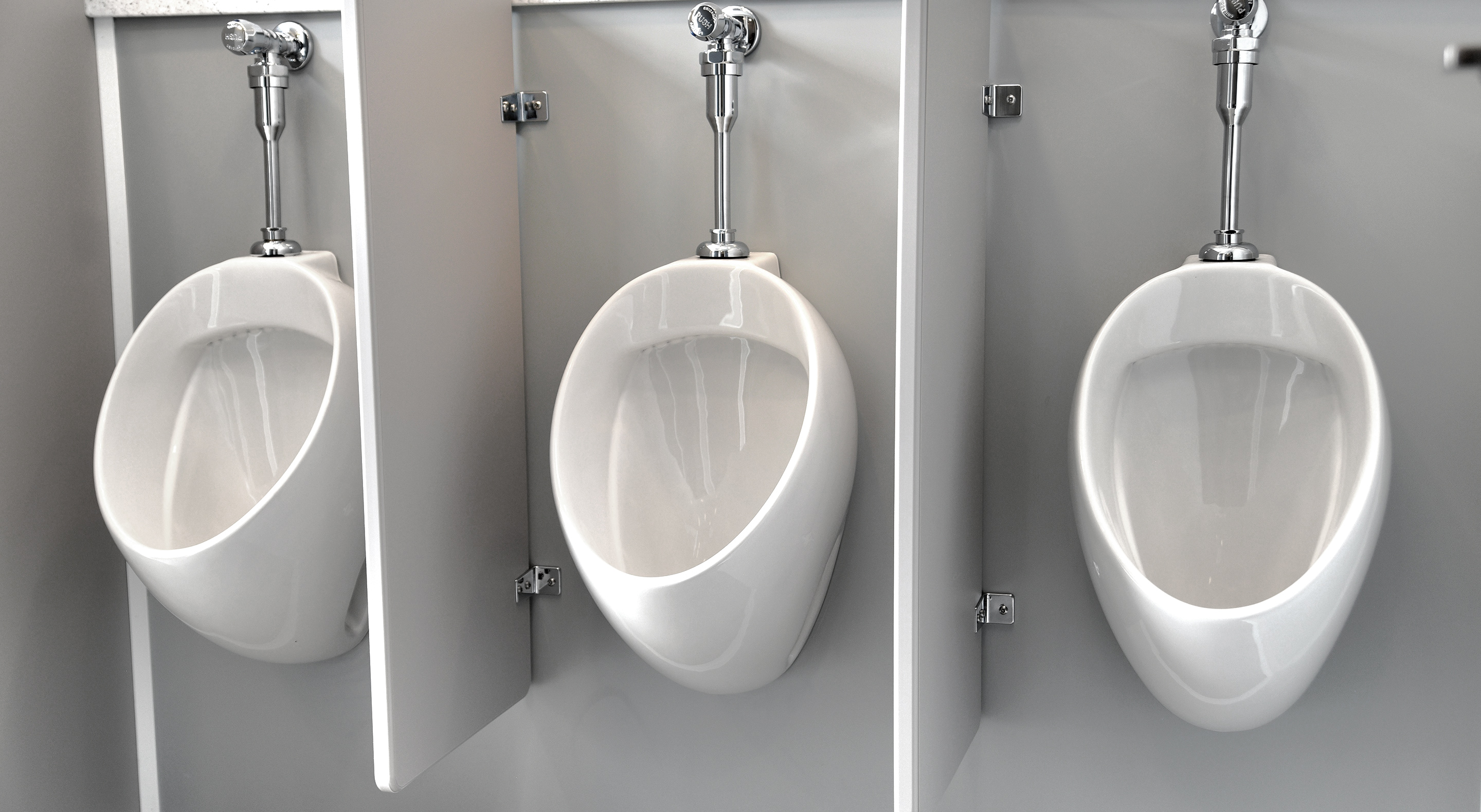Three white urinals mounted on a grey wall with dividers between them in a public restroom.