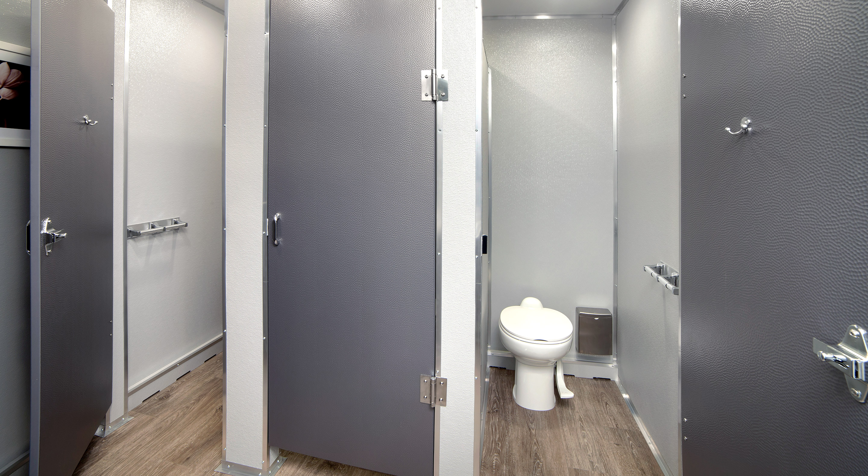 Interior of a clean public restroom with gray stalls and wood-style flooring, one open showing a toilet.
