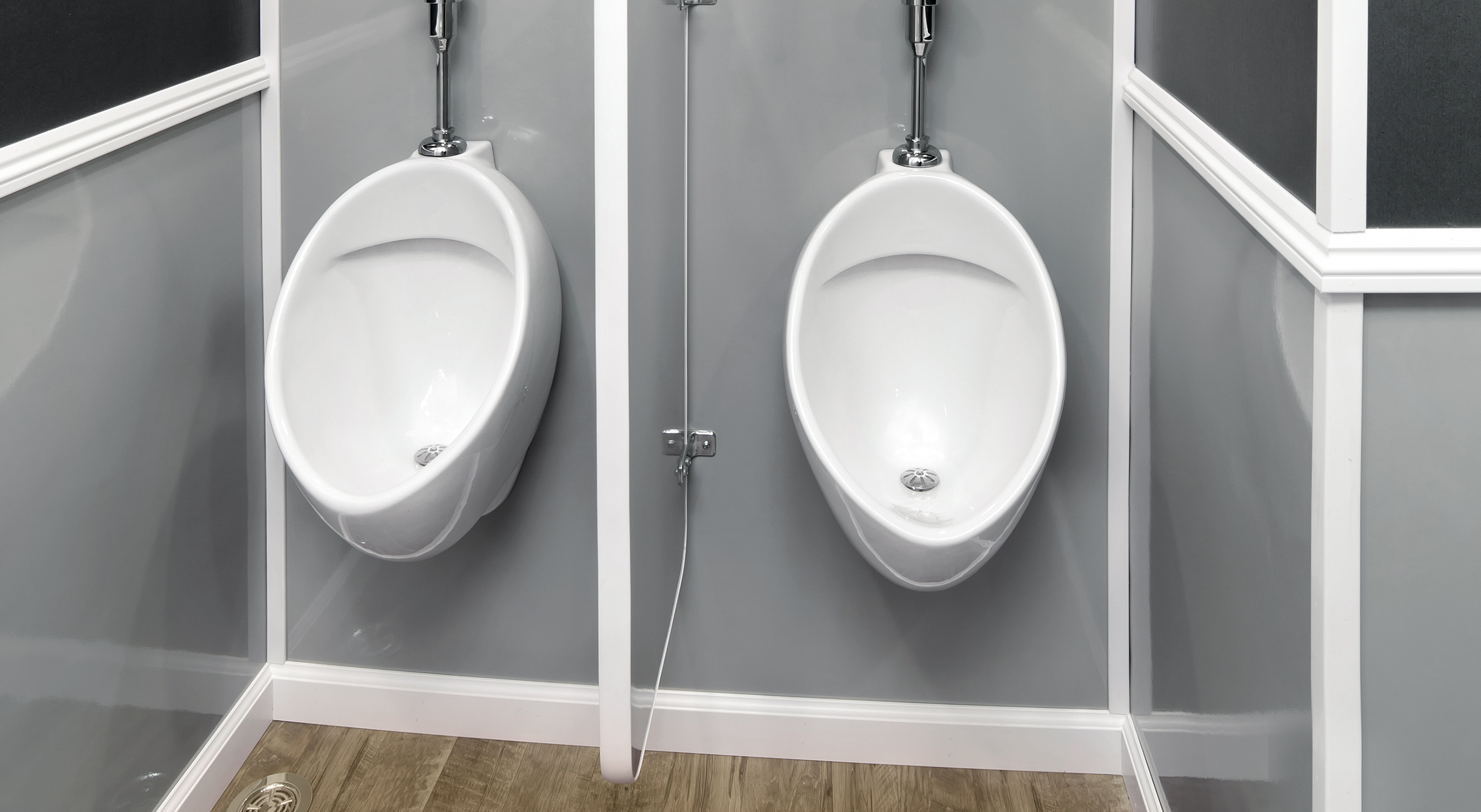 Two white urinals mounted on a grey wall, separated by a white partition. Wood-look flooring