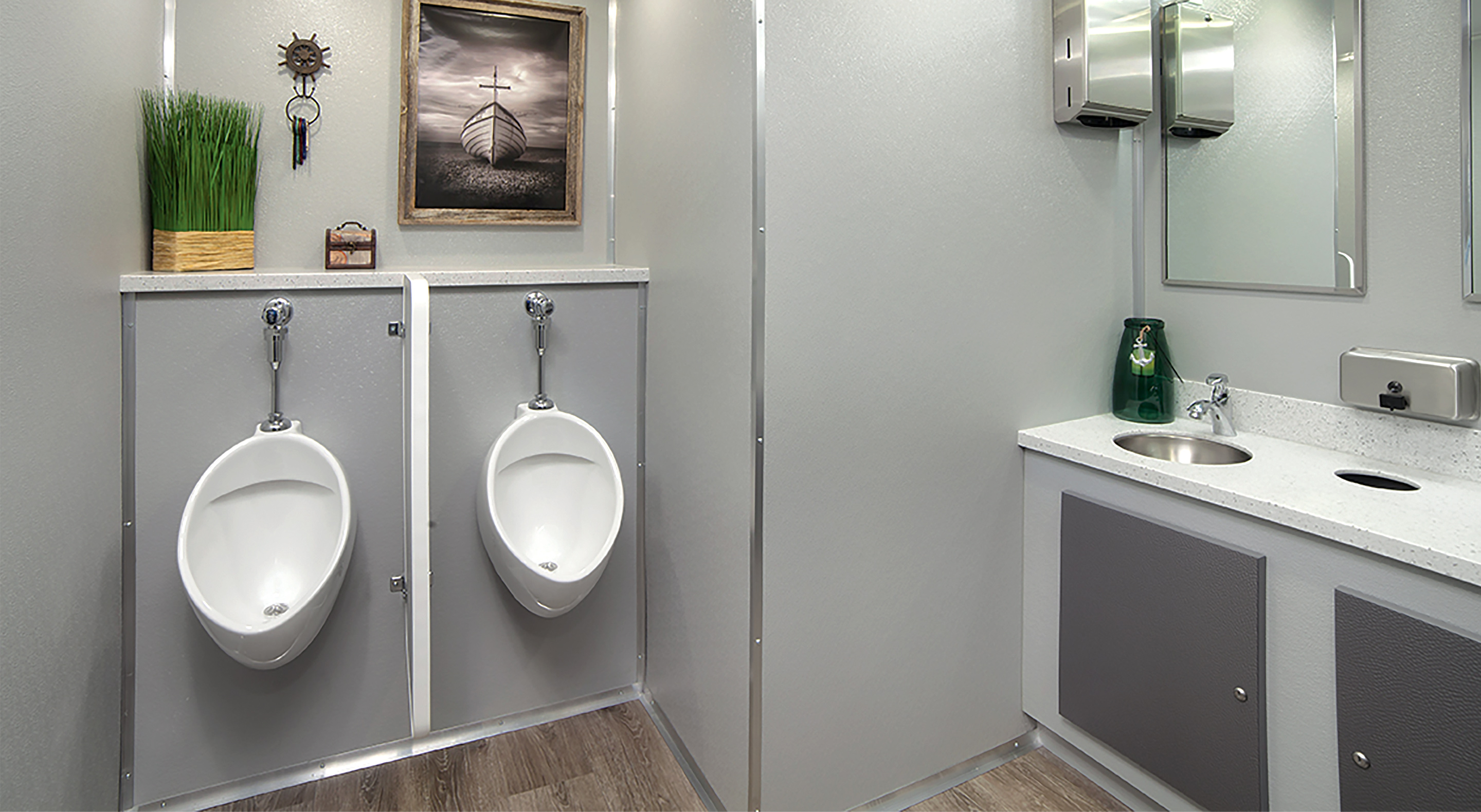 Modern restroom with two urinals, a sink, a mirror, green vase, grass plant, and framed boat photo.