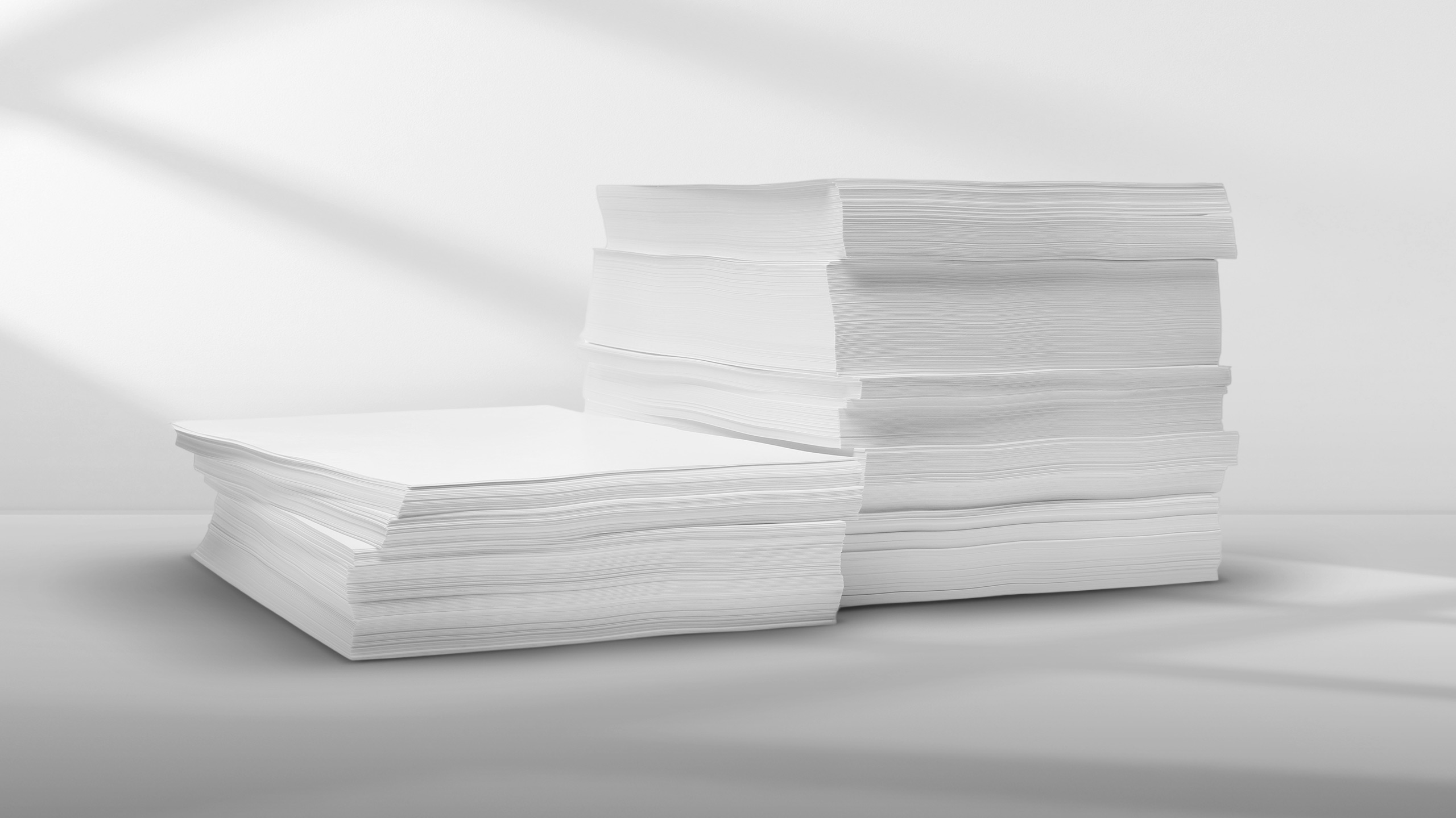 Two stacks of blank white printer paper on a smooth, light-gray surface with soft shadows.