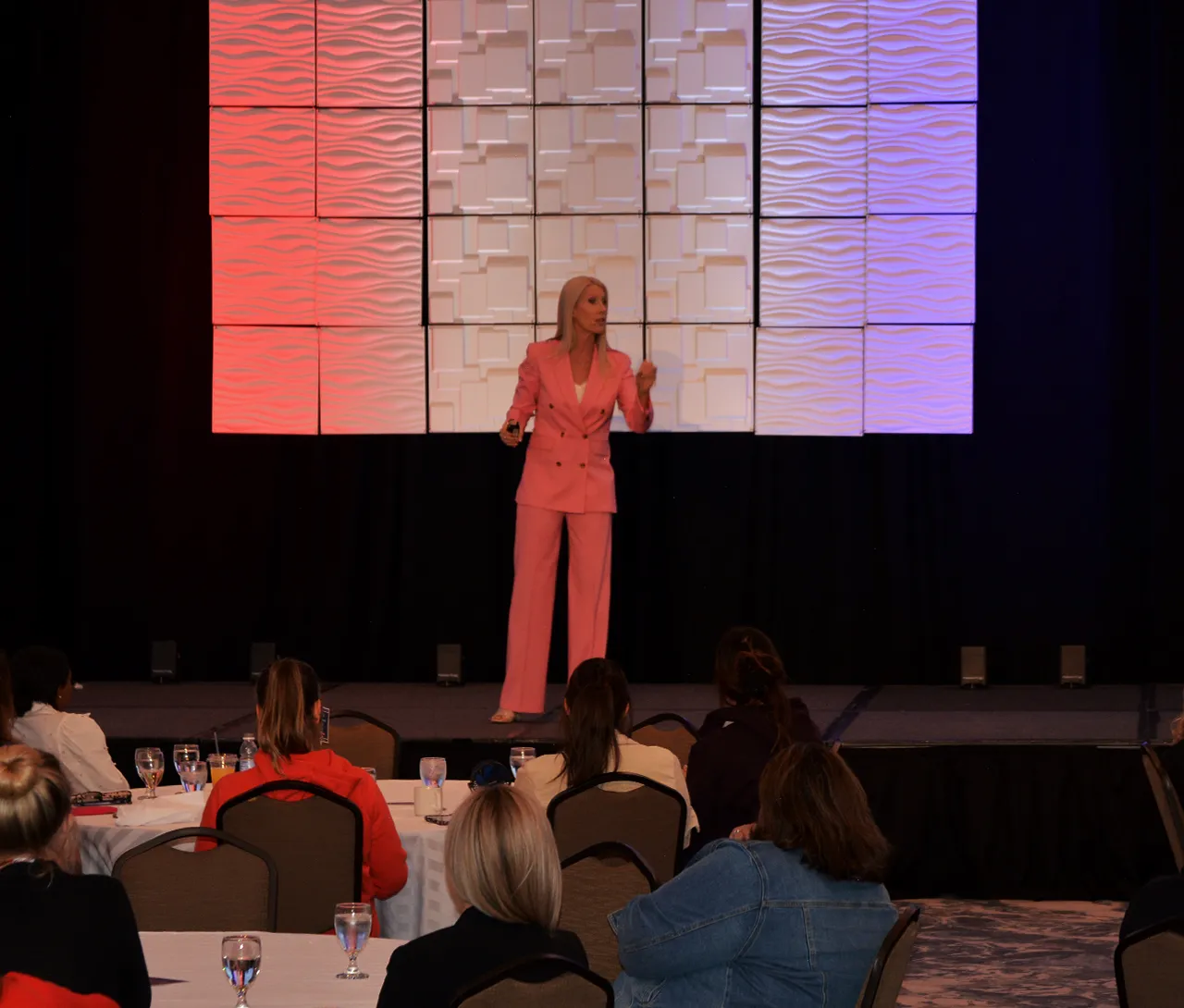 Woman in pink suit speaking on stage to seated audience with textured panel backdrop in red and blue lighting.