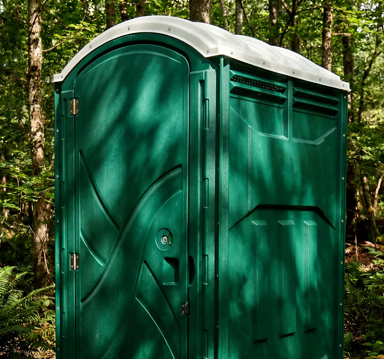 Green portable restroom with white roof standing outdoors among trees and foliage.