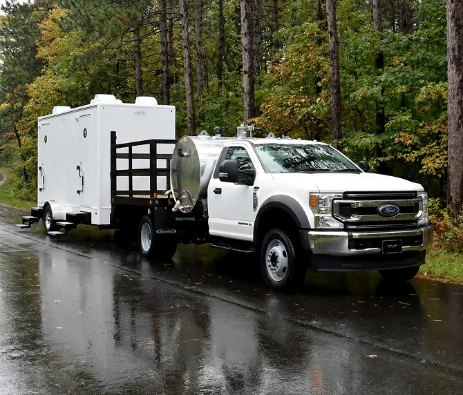White Ford pickup truck towing white trailer on wet forest road surrounded by tall trees.