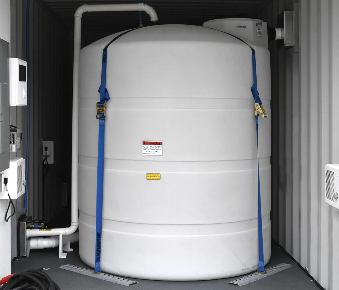 Large white cylindrical tank secured in a metal container with blue ratchet straps and connected pipes.