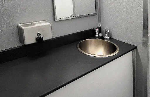 Small bathroom sink with stainless steel basin, faucets, black countertop, wall mirror, and soap dispenser.