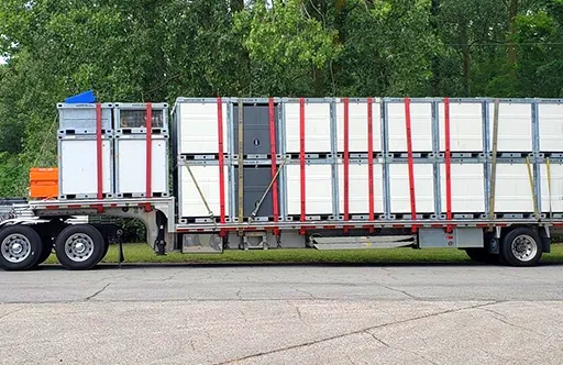 Truck trailer loaded with numerous white crates secured by red straps parked on pavement with trees behind.