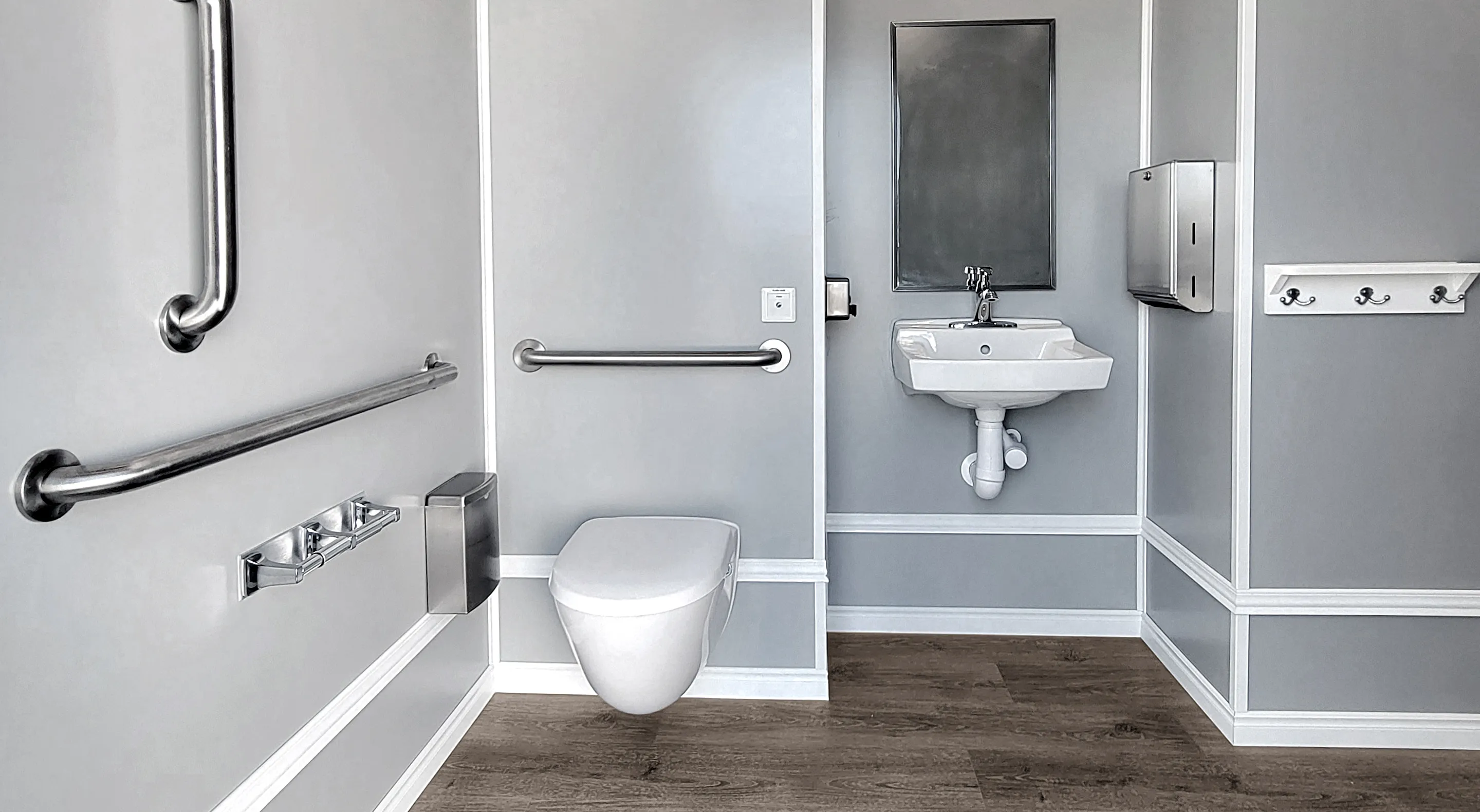 ADA-accessible bathroom with wall-mounted toilet, grab bars, sink, mirror, and hooks on gray walls.
