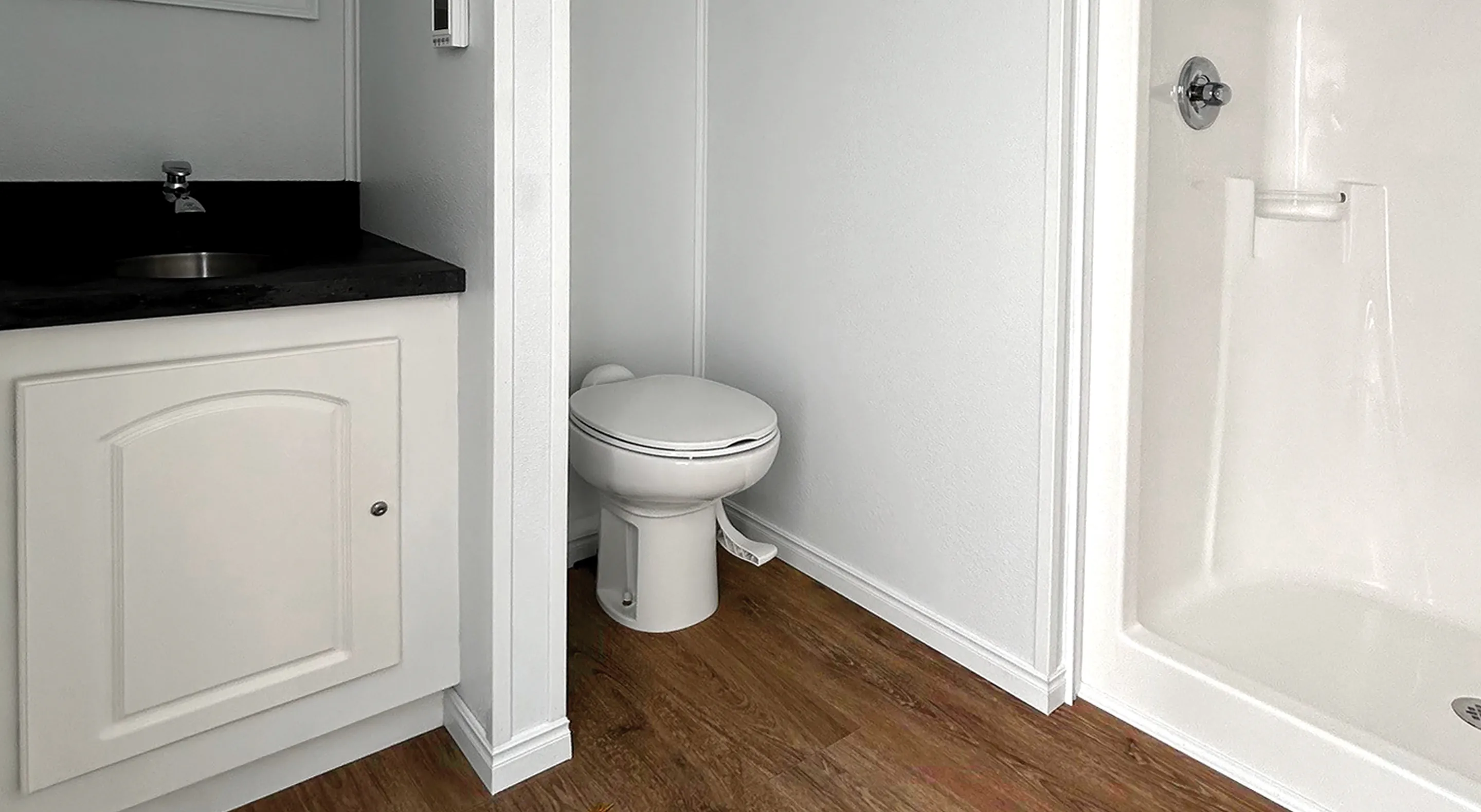 Small bathroom with white toilet, white shower, black countertop, and white cabinet over wood floor.